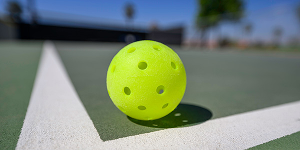 Close-up, selective focus shot of a bright neon-yellow pickleball sitting near the white corner lines of a green outdoor sports court.